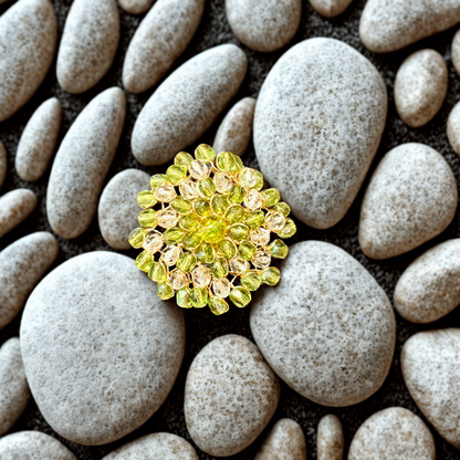 A round yellow-green floral brooch with faceted crystal-like petals, displayed on a bed of smooth gray river stones.