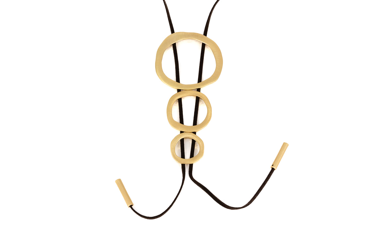 Three Circles Long Necklace with three graduated brushed gold Zamak circles threaded on dark Alcantara cords with gold-tipped ends, product shot on white background