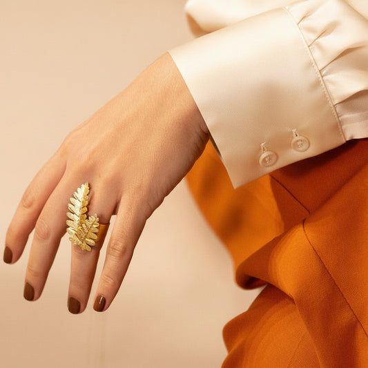 A close-up of a hand wearing a textured gold ring with a leaf or fern-like design, styled with a cream silk blouse and an orange garment against a warm beige background.
