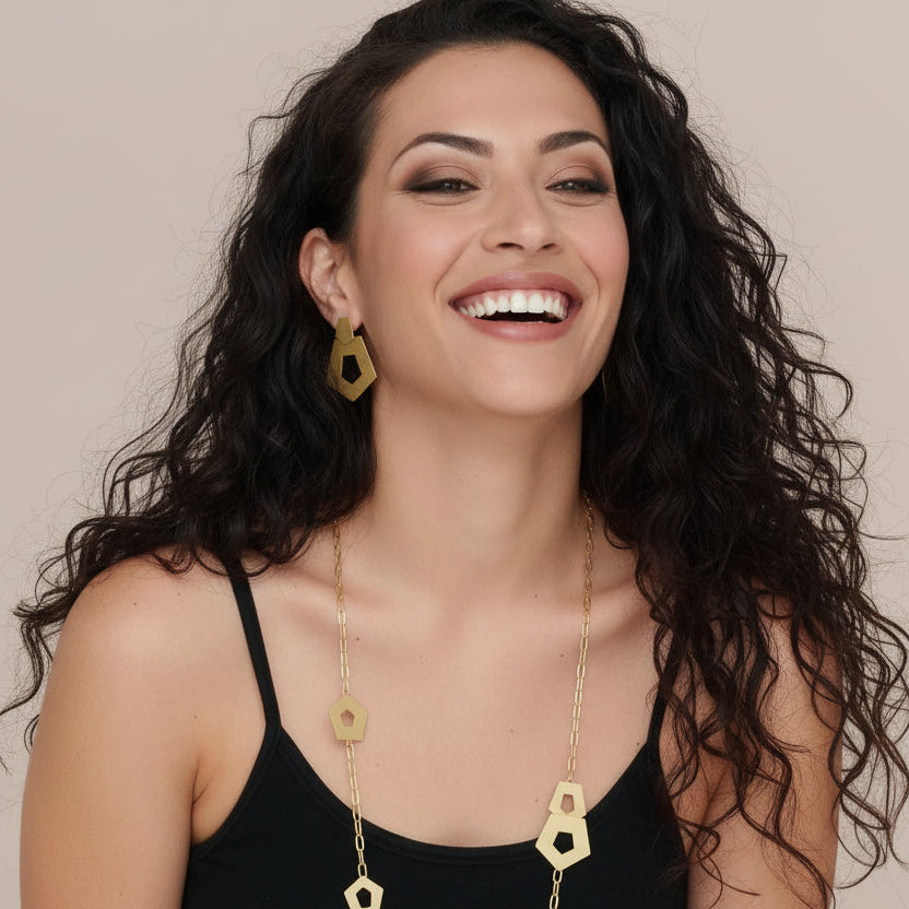 A woman with dark curly hair wearing a black camisole top models a gold geometric statement necklace and matching geometric drop earrings against a solid beige background.