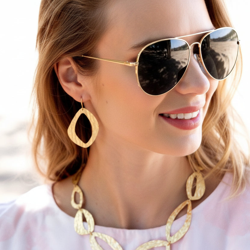 A close-up of a woman wearing a textured gold statement necklace and matching drop earrings, paired with a light pink top.
